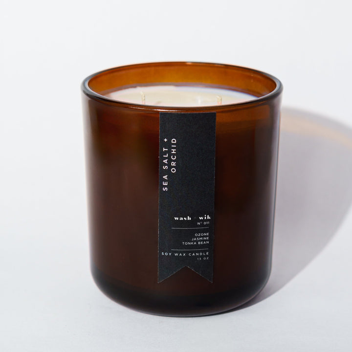 An amber glass jar candle with a black vertical label photographed on a clean white background with a soft shadow.