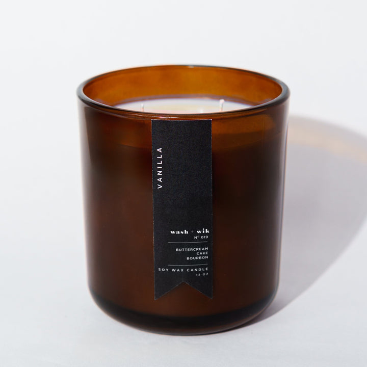 An amber glass jar candle with a black vertical label photographed on a clean white background with a soft shadow.