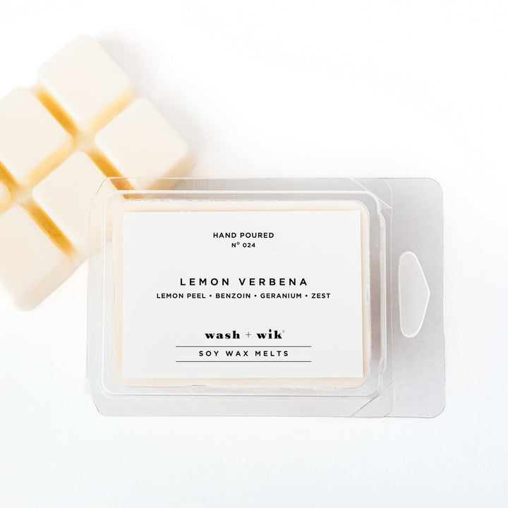 A clear plastic clamshell package of Wash + Wik soy wax melts on a white background, with several pale cream wax cubes partially visible behind it.