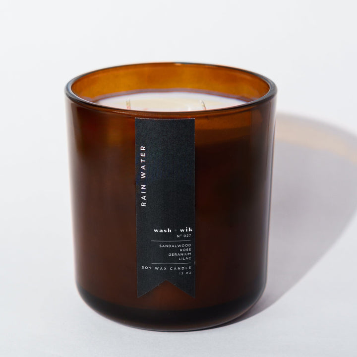 An amber glass jar candle with a black vertical label photographed on a clean white background with a soft shadow.