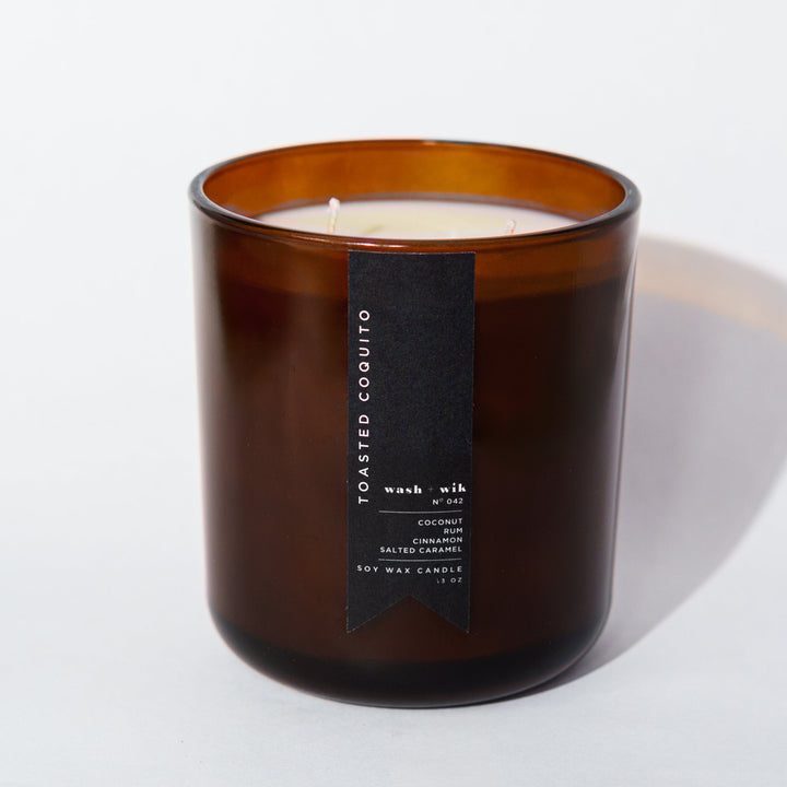 An amber glass jar candle with a black vertical label photographed on a clean white background with a soft shadow.