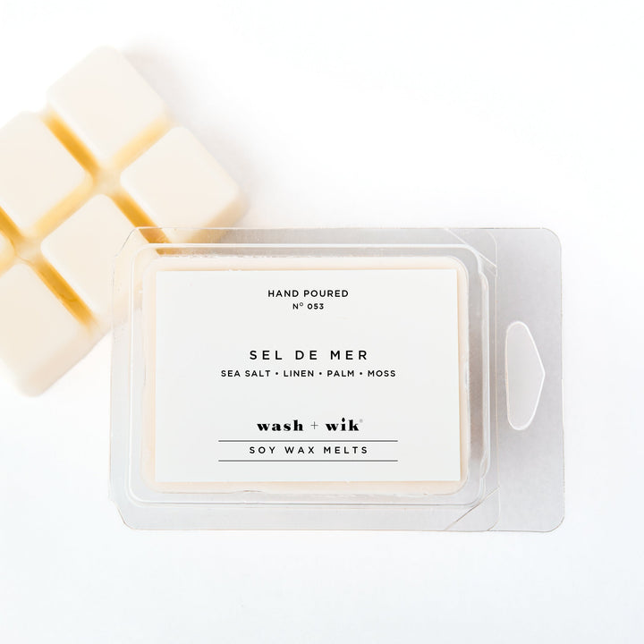 A clear plastic clamshell package of Wash + Wik soy wax melts on a white background, with several pale cream wax cubes partially visible behind it.
