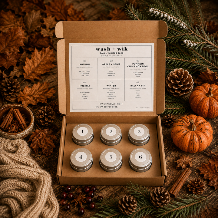 An open kraft cardboard box displaying six small round metal tins labeled 1 through 6, with a printed scent calendar card inside the lid. The box is styled on a rustic surface with autumn leaves, pinecones, cinnamon sticks, evergreen branches, and small pumpkins.