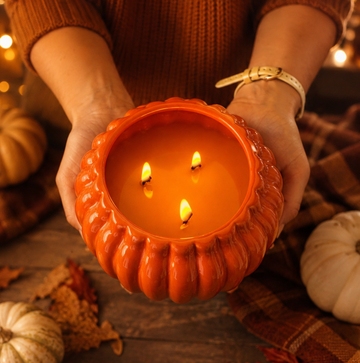 A person holds a glowing orange pumpkin-shaped candle with three lit wicks in both hands, surrounded by fall decor including pumpkins, a plaid blanket, and warm string lights.