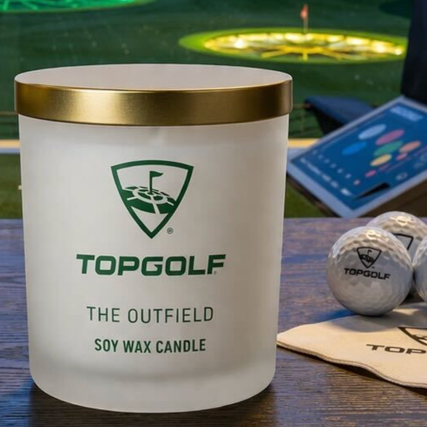 TopGolf candle with gold lid on a table with golf balls and a scorecard in the background.