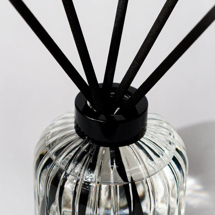 A close-up of a reed diffuser with black reeds inserted into a ribbed clear glass bottle with a black collar, set against a light gray background.