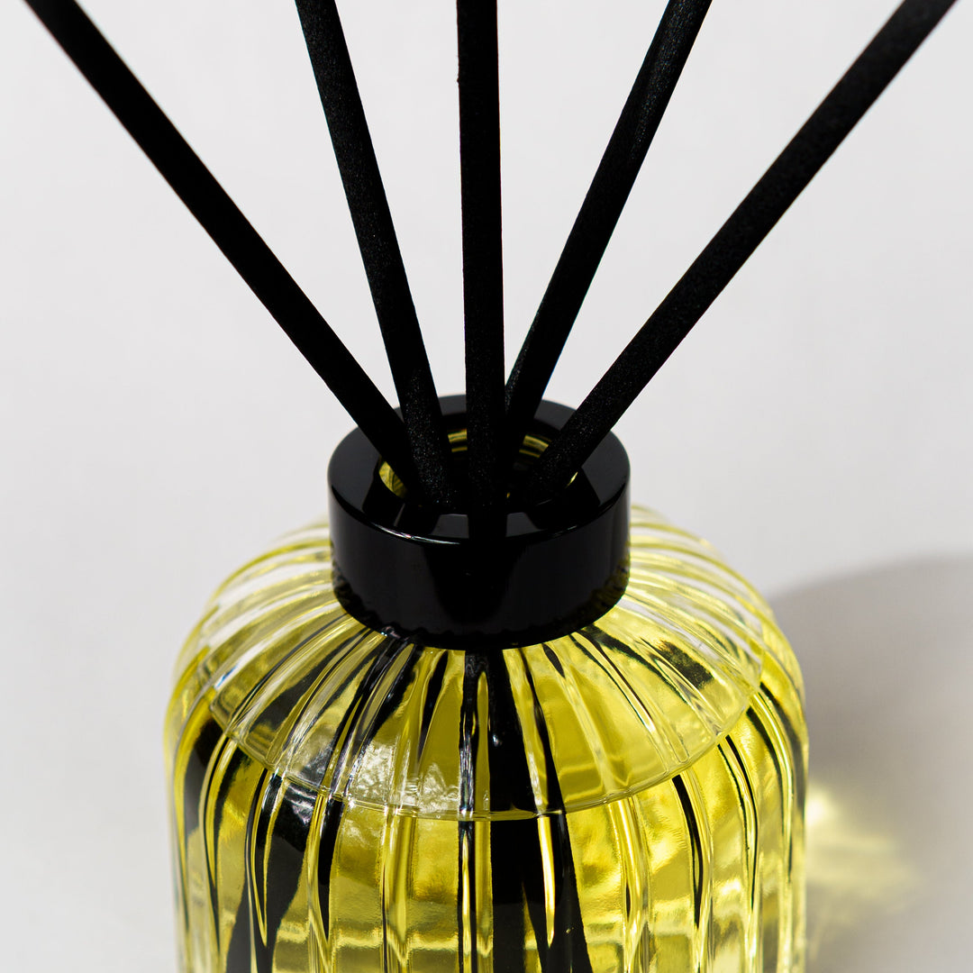 A close-up of a reed diffuser with a ribbed clear glass bottle filled with yellow liquid and black reeds extending upward against a light gray background.