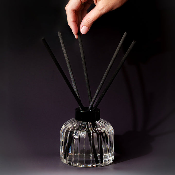 A hand is placing a black reed diffuser stick into a clear glass bottle with several black reeds, set against a dark studio background.