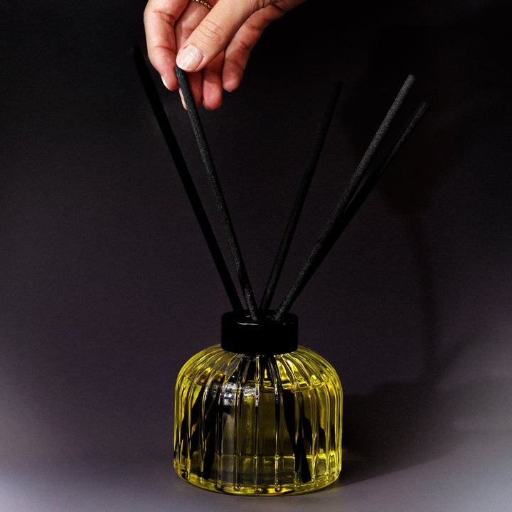 A hand is holding one of the black reed diffuser sticks above a ribbed glass bottle filled with yellow liquid, set against a dark studio background.