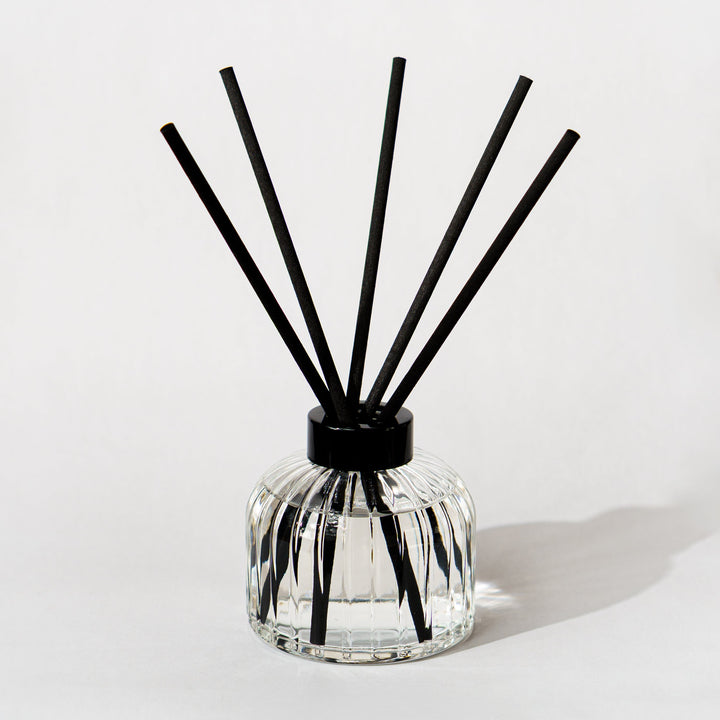 A reed diffuser with a faceted clear glass bottle and five black reeds, photographed on a clean white background.