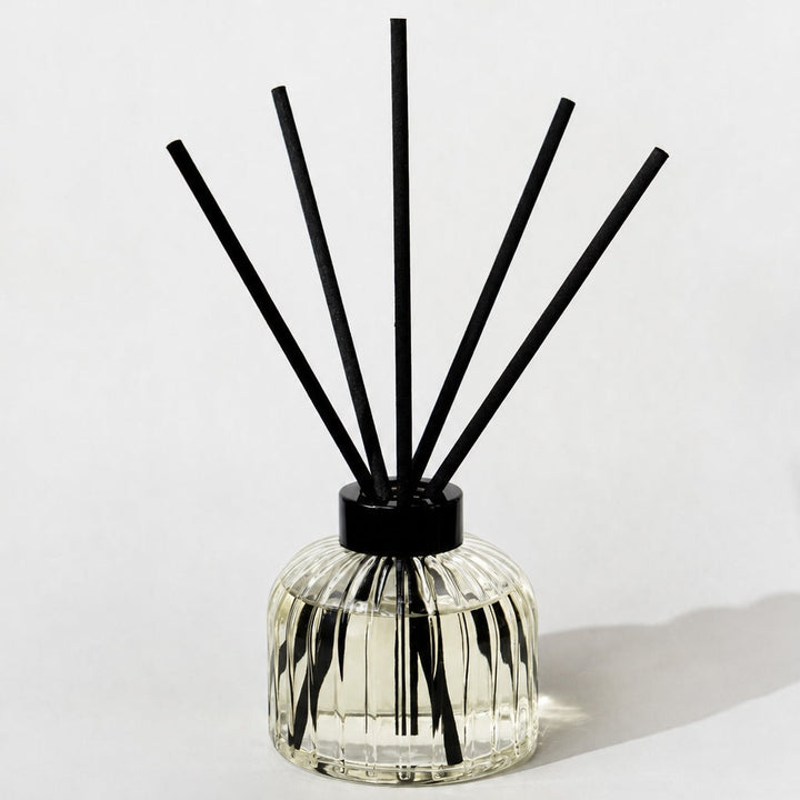 A clear glass reed diffuser bottle with a black cap and multiple black reeds, photographed on a clean white background with a soft shadow.