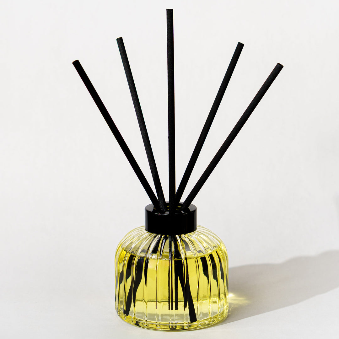 A reed diffuser with a ribbed clear glass bottle filled with yellow liquid, topped with a black cap and featuring five black reeds, set against a plain white background.