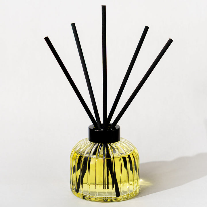A reed diffuser with a ribbed clear glass bottle filled with yellow liquid, topped with a black cap and featuring five black reeds, set against a plain white background.