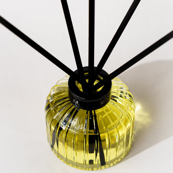 A reed diffuser with a ribbed translucent yellow glass bottle and multiple black reeds, photographed on a clean white background.
