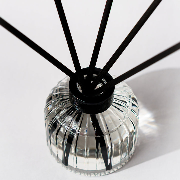 A reed diffuser with a clear ribbed glass bottle and black reeds, photographed on a clean white background.