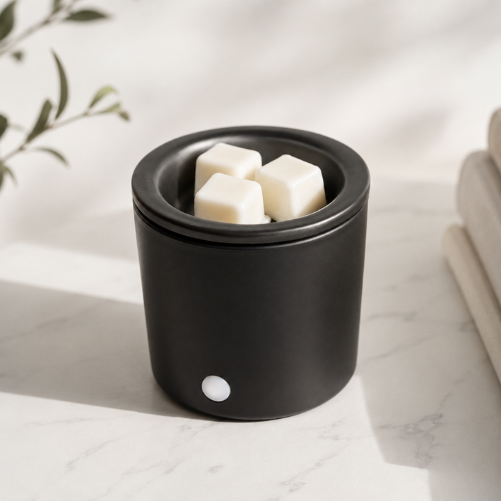 A matte black cylindrical wax warmer or melt pot with three off-white wax cubes inside, placed on a light marble surface with soft natural shadows.