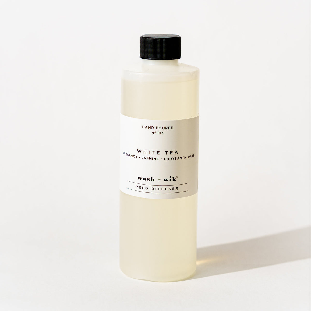 A bottle of Wash + Wik reed diffuser refill with a black cap and a minimalist white label, set against a clean white background with a soft shadow.