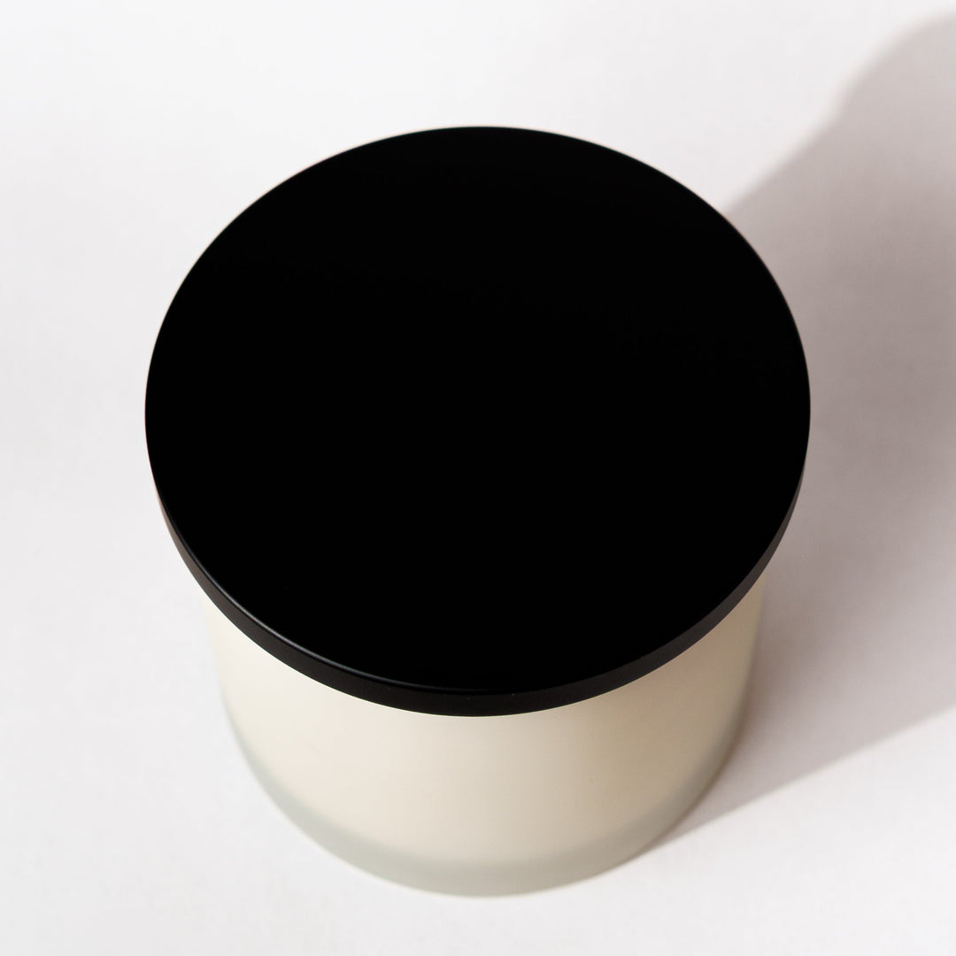 A minimalist cylindrical container with a matte black circular lid and a frosted off-white body, photographed from a top-down angle on a clean white background with a soft shadow.